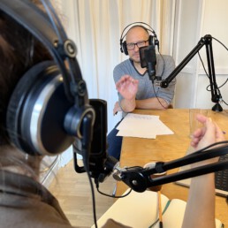 Zwei Personen führen ein Podcast-Interview im Studio; Kopfhörer, Mikrofonarme mit Popfilter, Notizblätter und Glas auf Holztisch; über-die-Schulter-Perspektive.