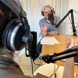 Zwei Personen führen ein Podcast-Interview im Studio; Kopfhörer, Mikrofonarme mit Popfilter, Notizblätter und Glas auf Holztisch; über-die-Schulter-Perspektive.