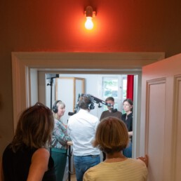 Blick durch eine geöffnete Tür in ein Podcaststudio; rote On‑Air‑Lampe signalisiert Aufnahme; mehrere Personen mit Kopfhörern sprechen an Mikrofonen; Fenster und roter Vorhang im Hintergrund.