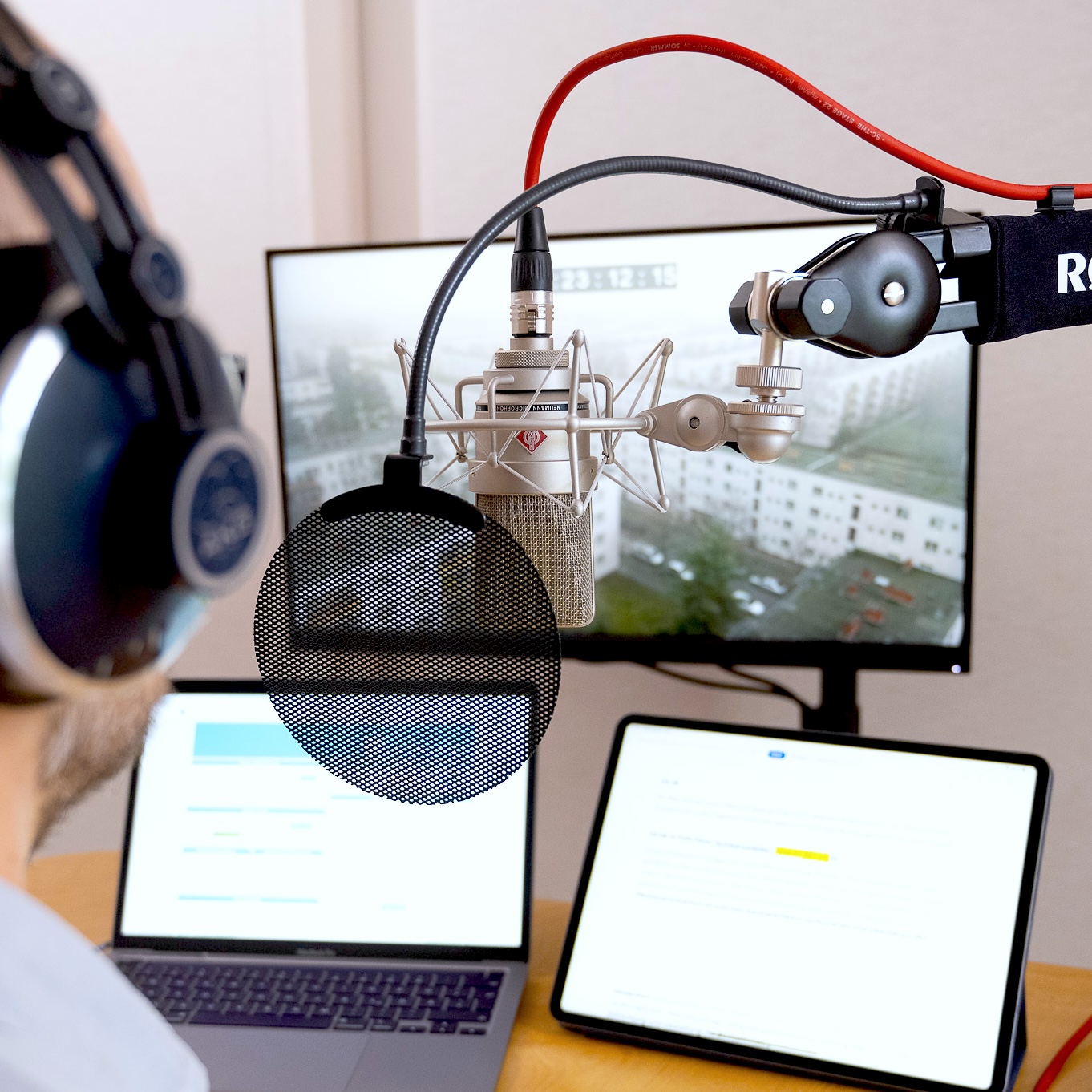 Person mit Kopfhörern spricht in ein Neumann Großmembran‑Mikrofon mit Popfilter am Mikrofonarm; Laptop und Tablet auf dem Tisch; Monitor im Hintergrund.
