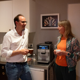 Zwei Personen unterhalten sich an der Studioküche und trinken Kaffee; Vollautomat auf der Arbeitsfläche; lockere Gesprächsszene vor/zwischen Podcastaufnahmen.