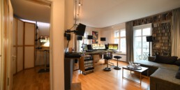 Helles Tonstudio mit Regieraum (Mischpult, Monitore, Lautsprecher), separatem Aufnahmeraum links und Lounge mit Sofa und Fensterfront.